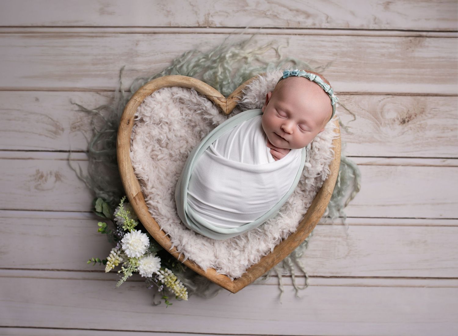 Baby girl wrapped in pale green and white, placed gently in a wooden heart prop during newborn studio photoshoot Renfrewshire