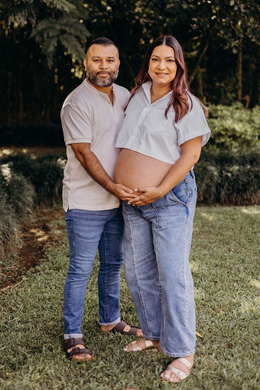 A couple in matching white shirts and jeans embrace while standing on grass in an outdoor photoshoot.