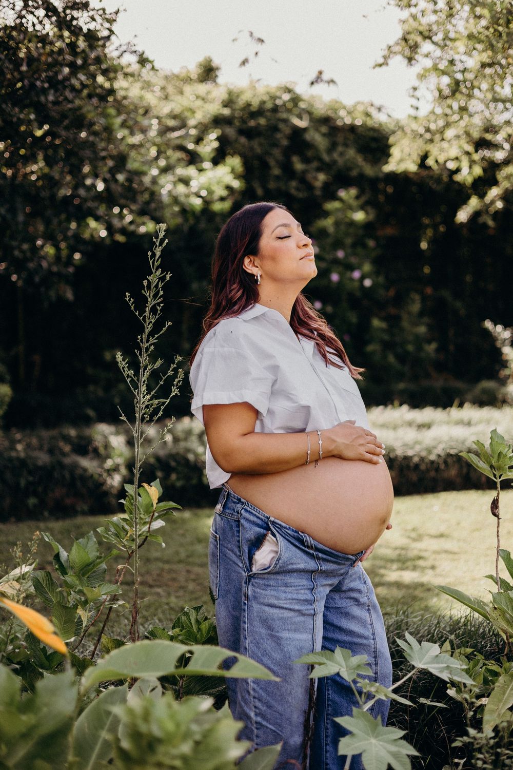 Maternity photoshoot in a garden setting with white shirt and denim overalls showing baby bump.