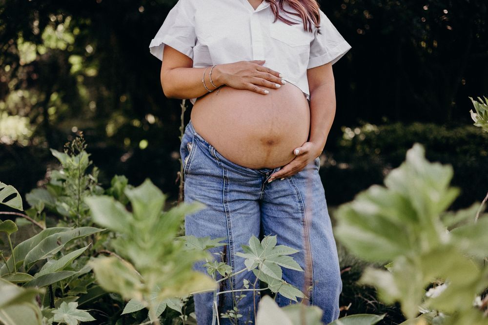 A pregnant person in blue jeans and white t-shirt stands in a garden with large green leaves in the background.