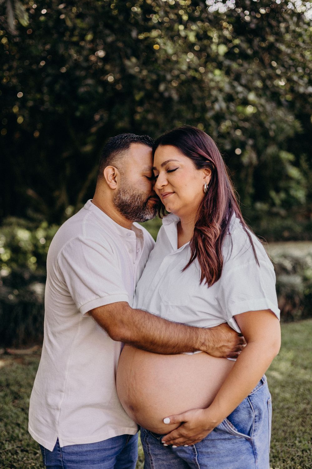 Expecting couple embraces tenderly in white shirts during outdoor maternity photoshoot.