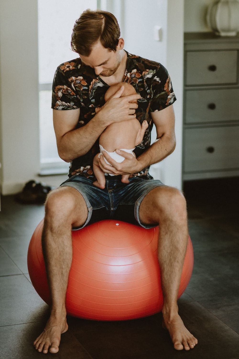Pappa sitter på en röd yoga boll och har sin nyfödda bebis i famnen. Bebisen har blöja. Dom sitter i hallen under en hemma-hos fotografering