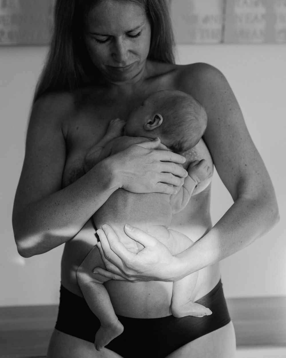 Svartvit bild på nyfödd bebis och hans mamma. Mamma och bebis har bar hud och mamman håller om bebisen med båda sina händer.Dom står i köket