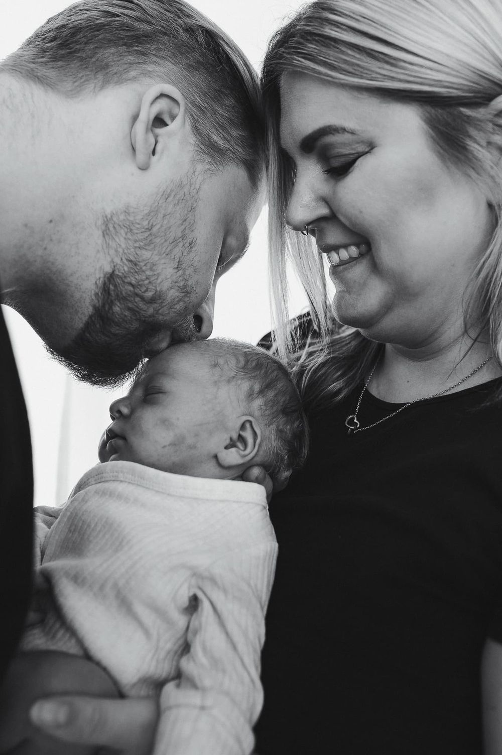 Nyfödd bebis ligger mellan mamma och pappa. Pappan pussar bebisen på huvudet. Bilden är svartvit.