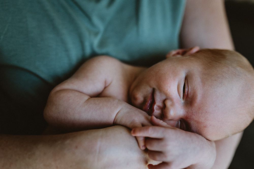 Nyfödd bebis ligger i sin mammas famn och det faller in naturligt ljus från ett fönster. Bebisen ler.