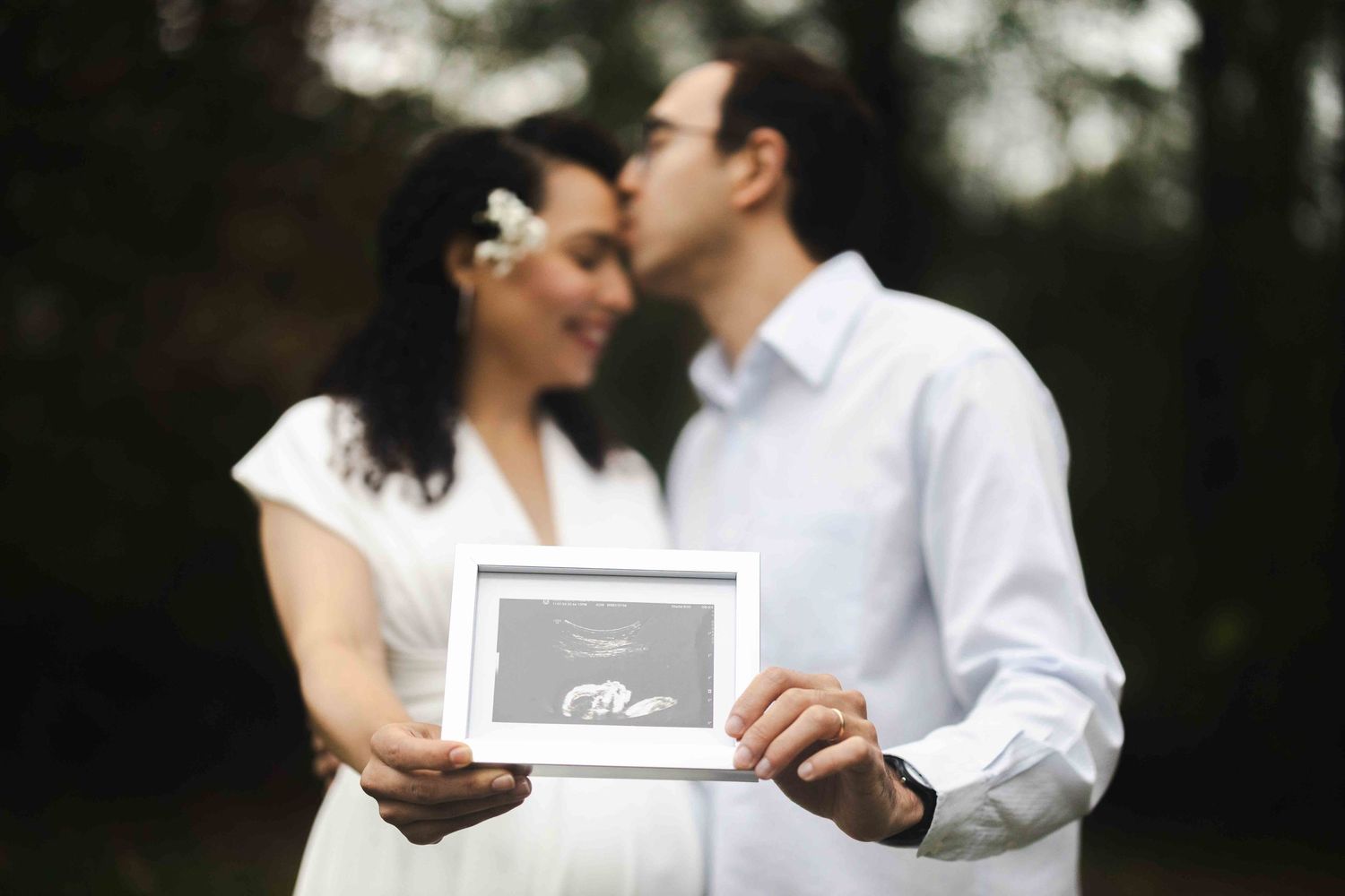 A couple shares an intimate moment while holding a framed ultrasound photo in an outdoor setting.