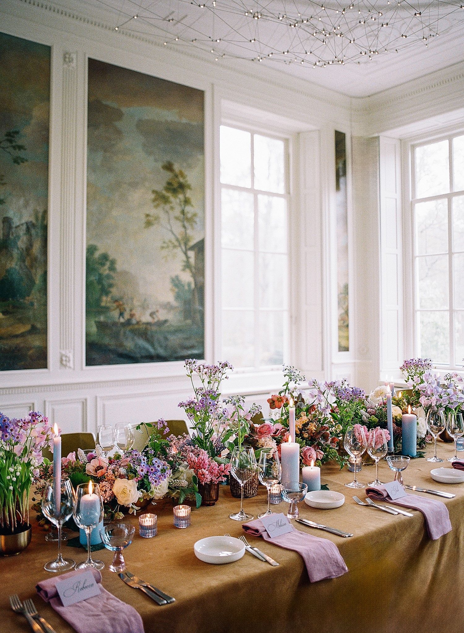 Elegant dining room with ornate ceiling, wall murals, and long table decorated with colorful floral arrangements and purple linens.