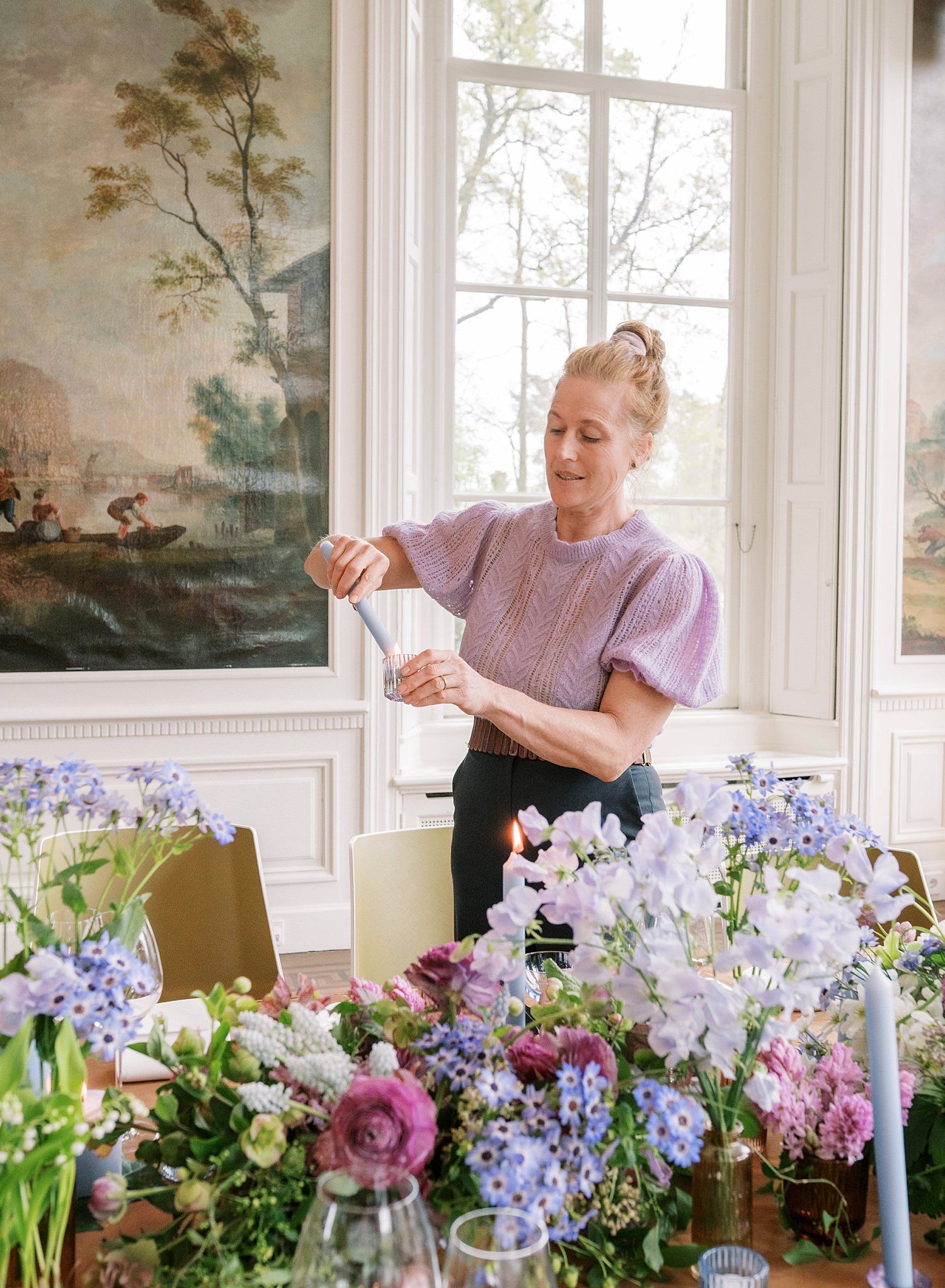 Person arranges fresh cut flowers in vases by a bright window with delicate purple and pink blooms on display.