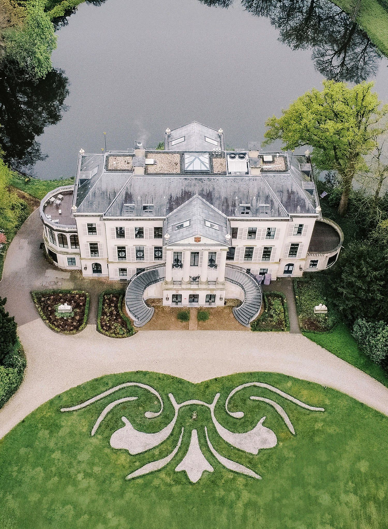 Aerial view of a luxurious white mansion with curved architecture and ornate landscaping featuring symmetrical garden designs.