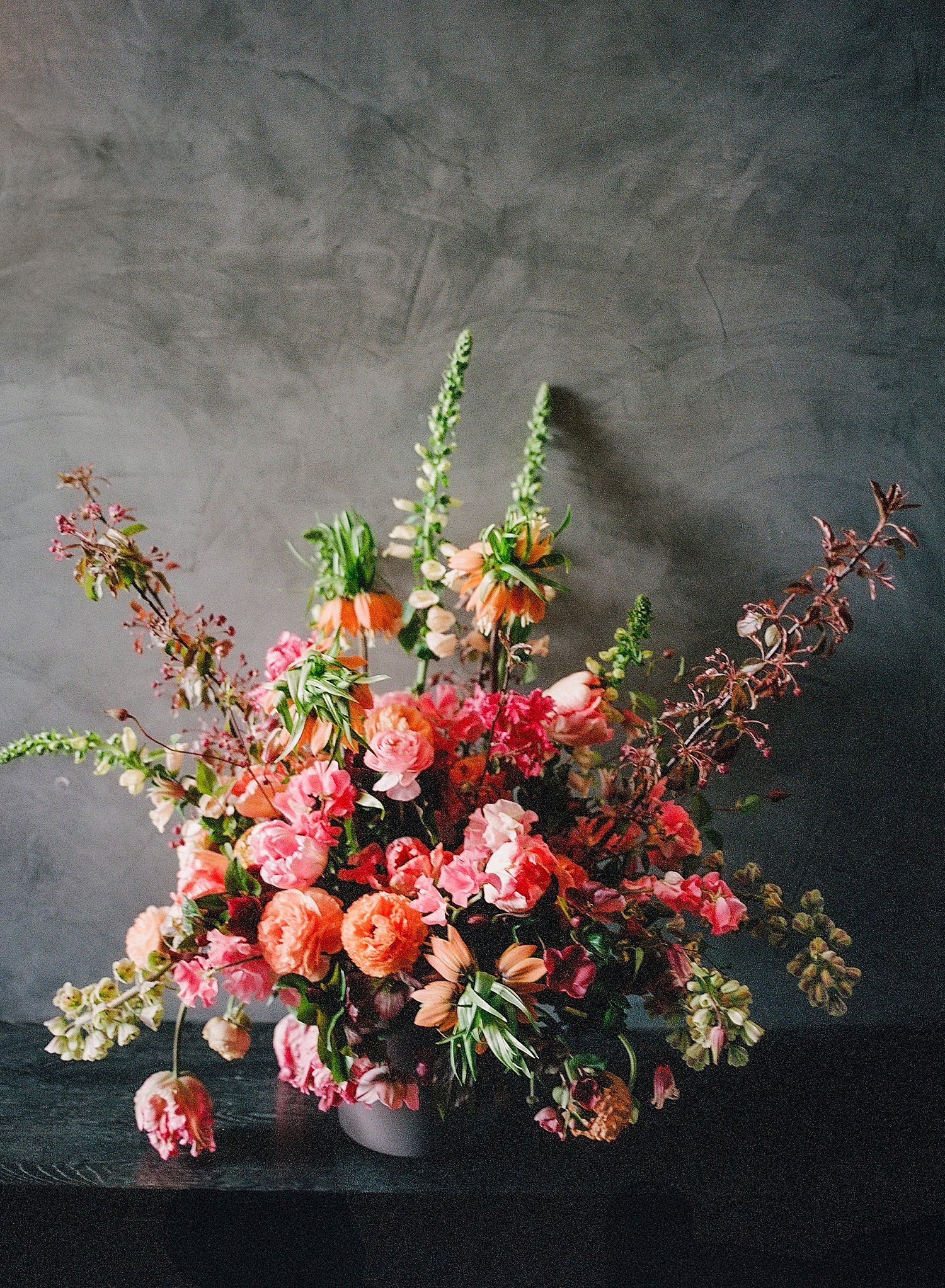 Lush floral arrangement with coral peonies, pink roses and delicate greenery against a moody dark gray background.