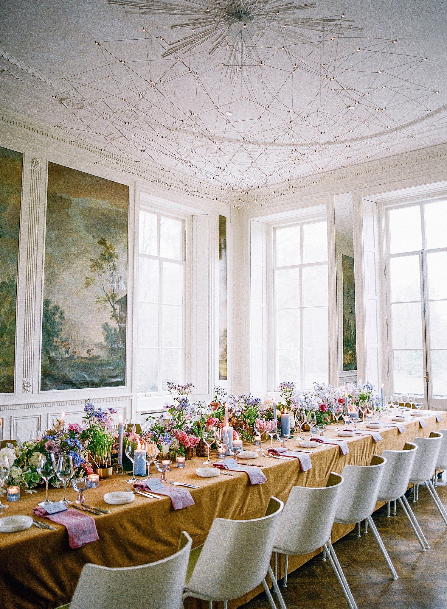 Elegant banquet hall with ornate ceiling details, white walls, and long tables decorated with colorful floral arrangements.