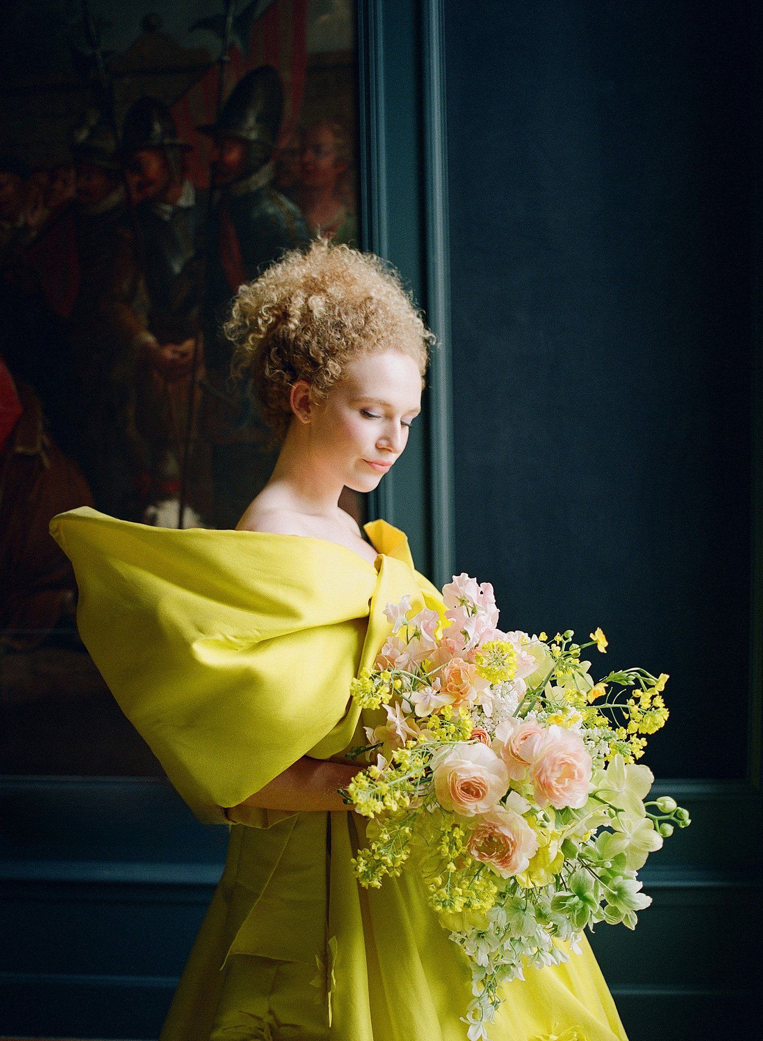 A figure in a flowing yellow dress holds a delicate bouquet of pink and white flowers against a dark background.