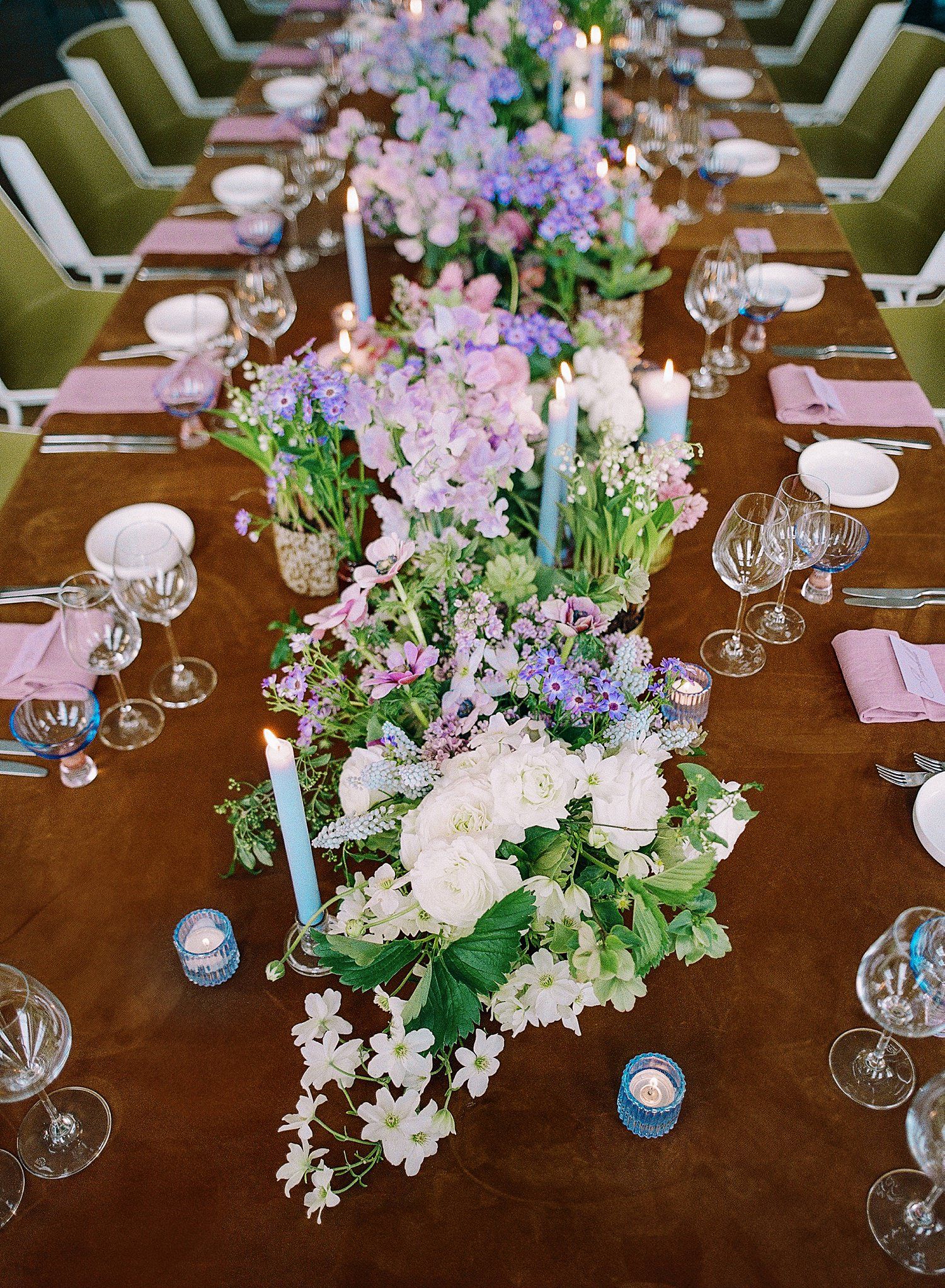 Elegant wedding table setting with pastel purple and white floral centerpieces, candles and crystal glassware on wooden table.