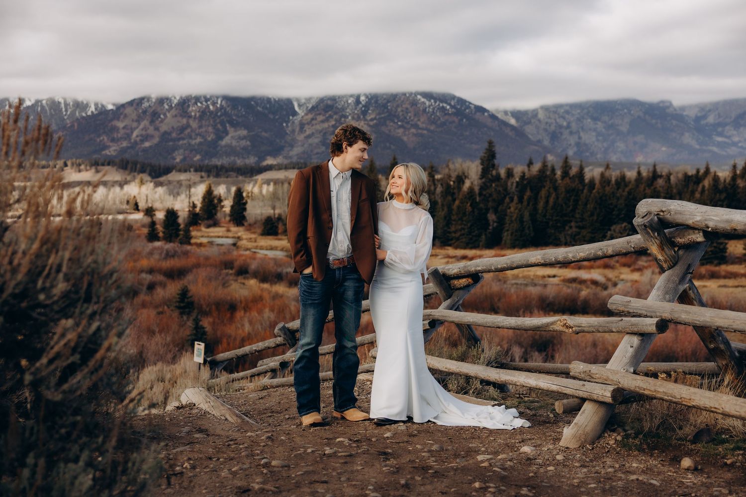 A couple in wedding attire stands on a scenic mountain overlook with snow-capped peaks in the distance.