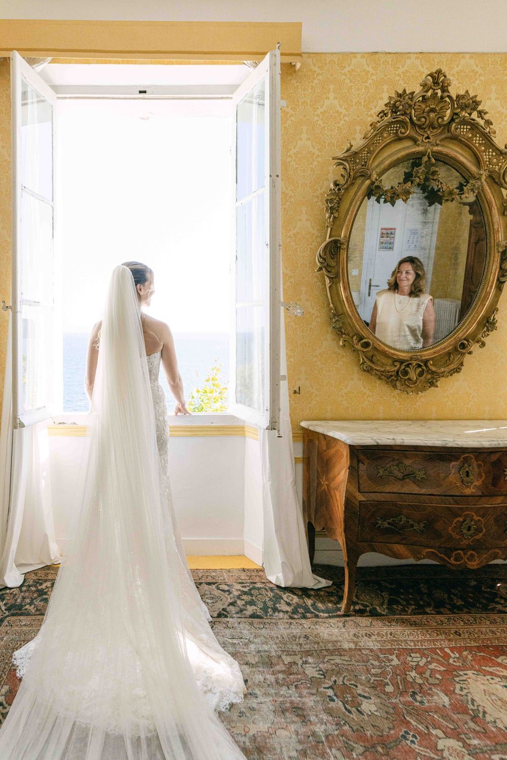 Mariée de dos devant une grande fenêtre, longue traîne et voile, miroir doré, lumière naturelle en intérieur.