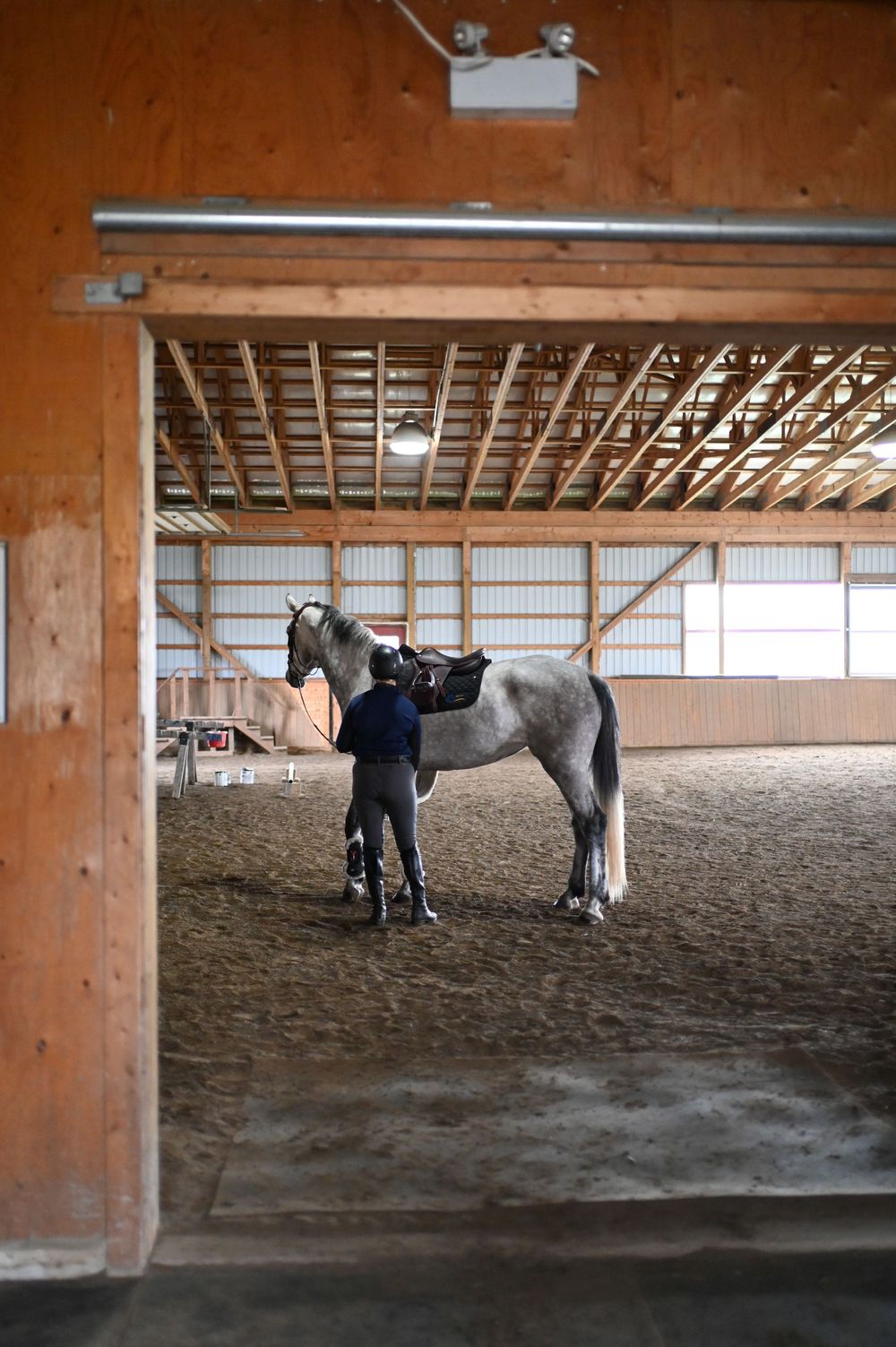 People working with horses in an indoor riding arena with wooden beams and sandy ground.