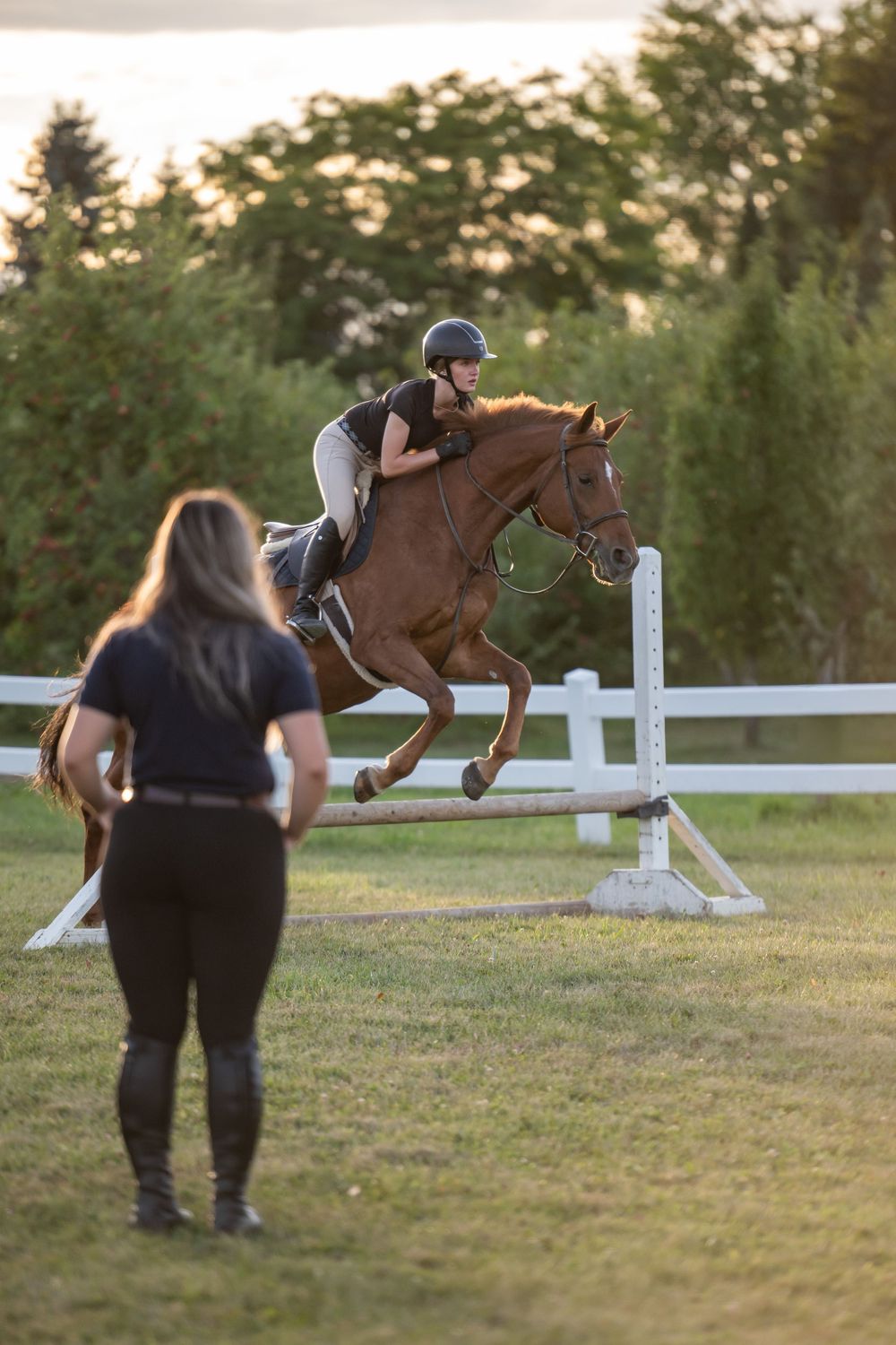 A rider and horse leap over a white jump rail while an instructor observes during an outdoor equestrian training session.