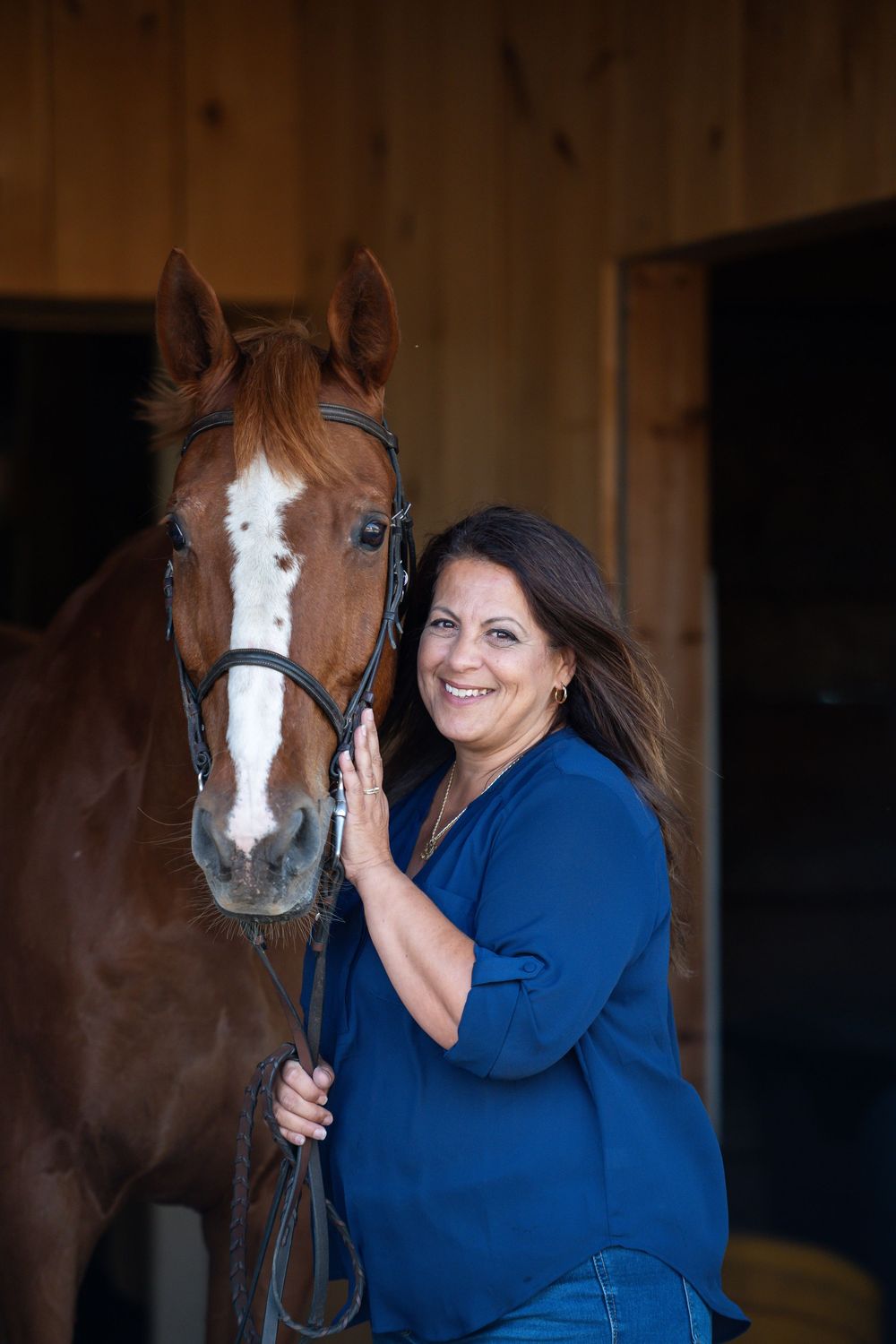 A woman in a blue sweater shares affectionate moments with a brown and white horse in a barn setting.