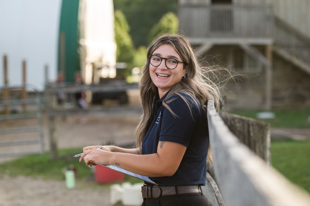 A woman in a black polo shirt smiles while standing outdoors near a building.