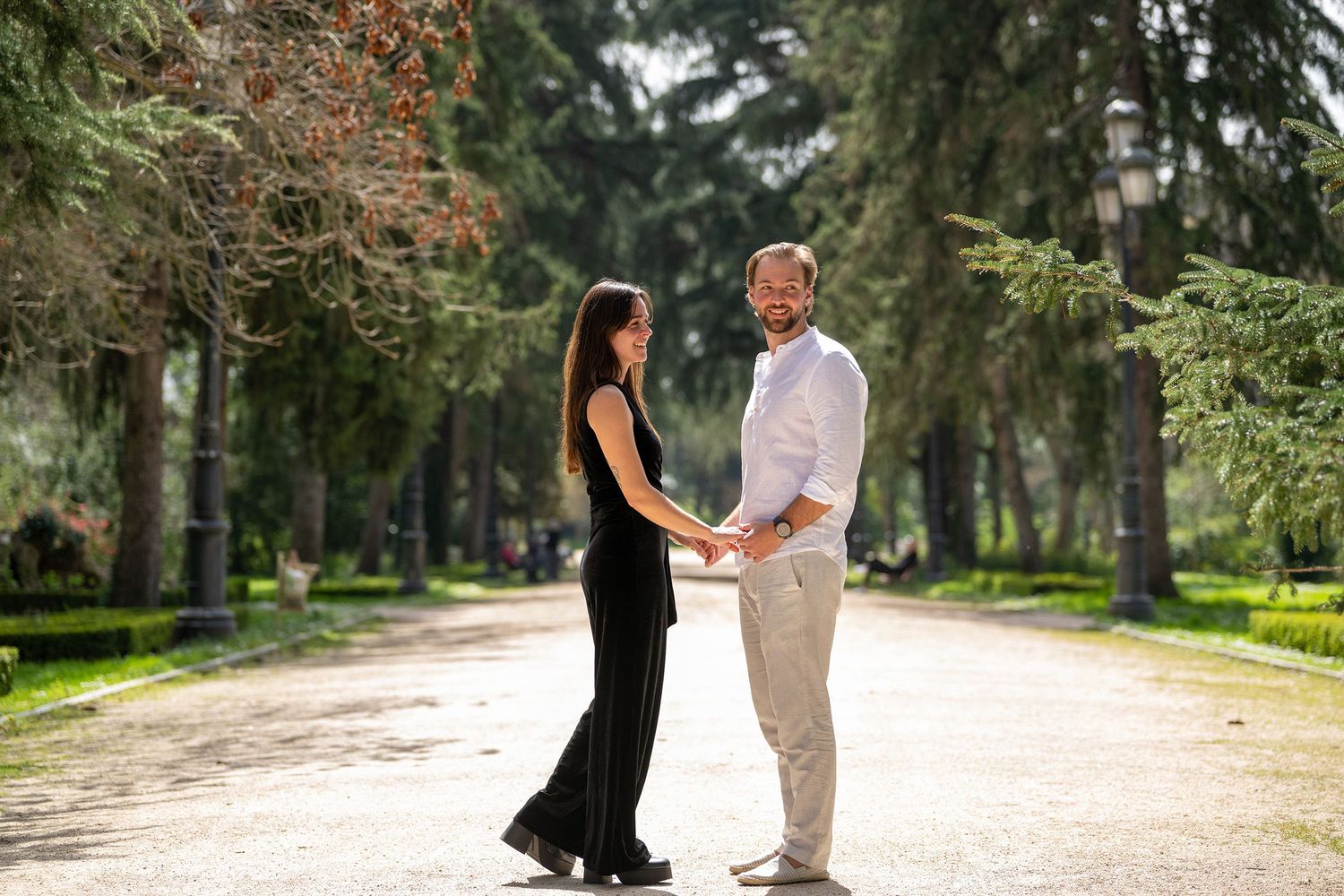 A couple stands together on a tree-lined path in a romantic outdoor setting during a photo session.
