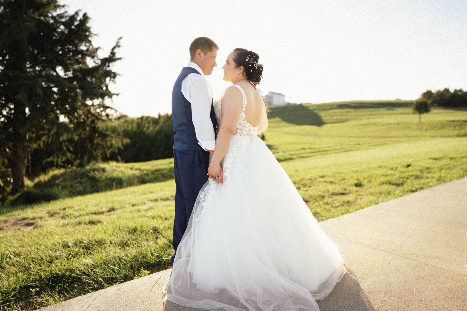 Newlyweds in formal attire embrace on path with rolling green hills in background.
