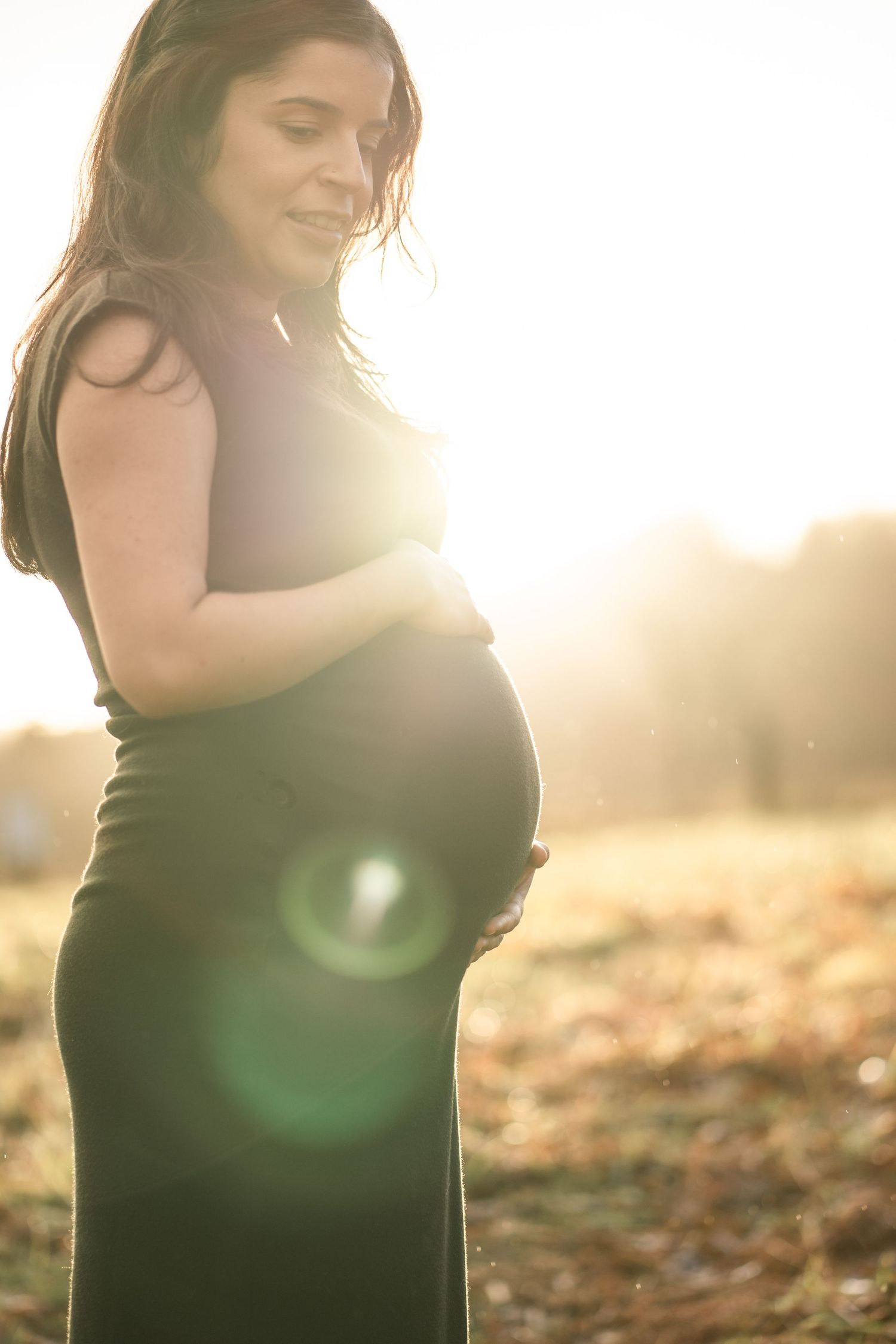 A woman in a black dress stands in golden sunlight among autumn leaves during a maternity photoshoot.