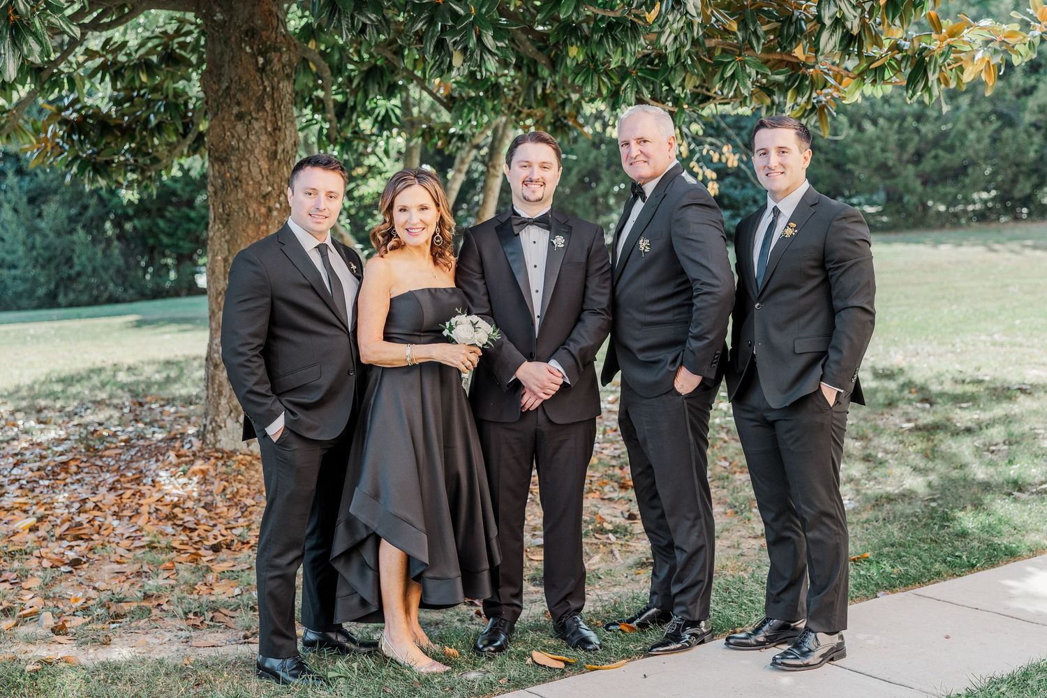 Wedding party in formal attire poses together outdoors on stone pathway surrounded by autumn foliage.