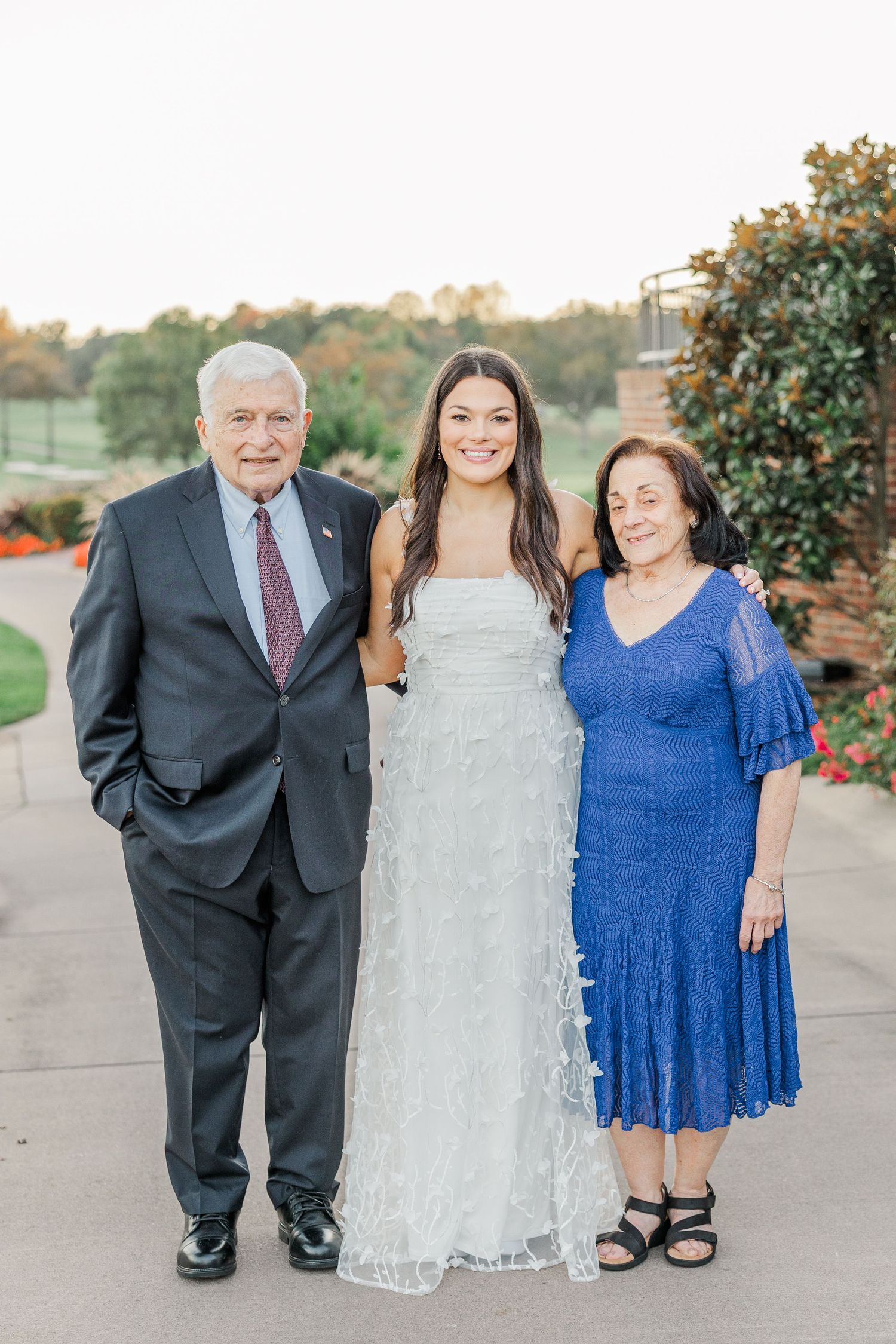 Three people pose together outdoors wearing formal attire including a blue dress and white lace gown.