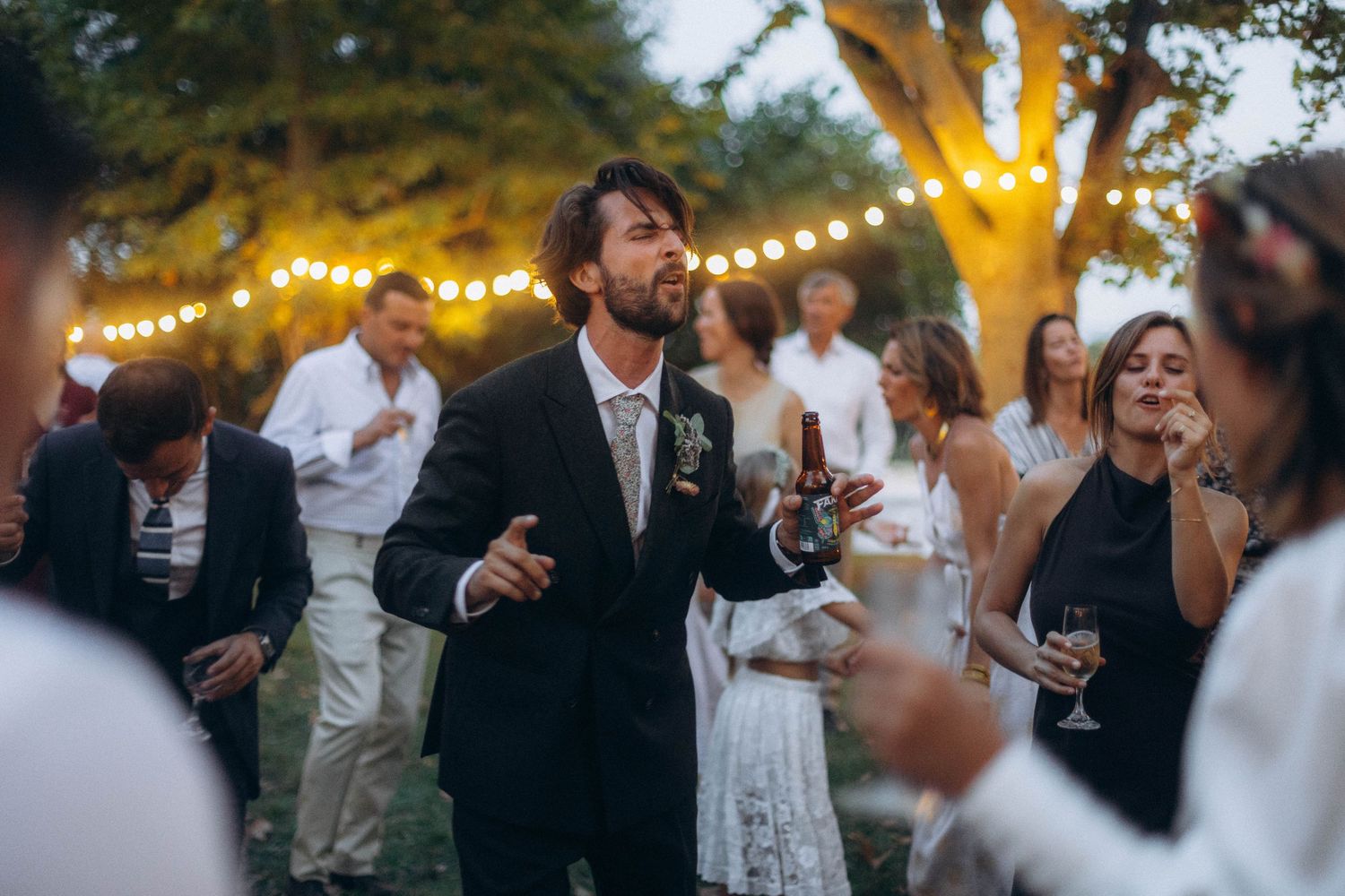 Guests gather outdoors at dusk for an elegant garden party celebration with string lights illuminating the trees.