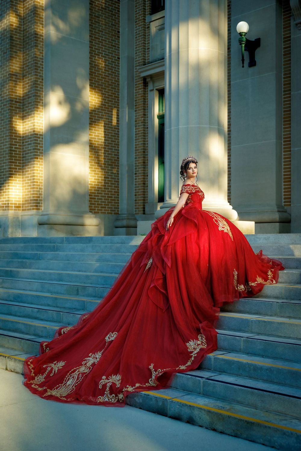 Woman in stunning red and gold embroidered ball gown poses gracefully on classical building steps.