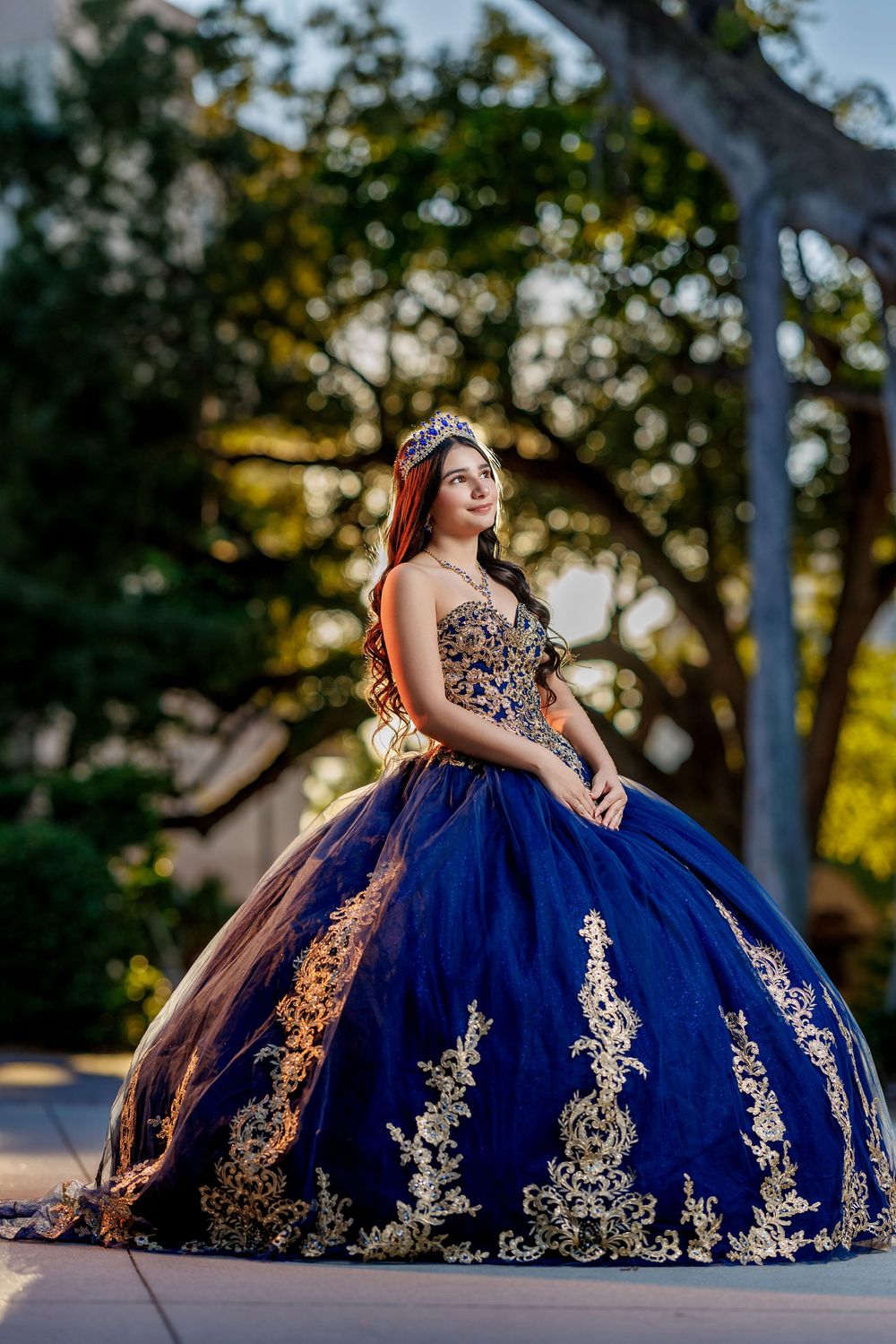 Young woman in navy blue quinceañera ball gown with gold embroidery and tiara poses outdoors at sunset.