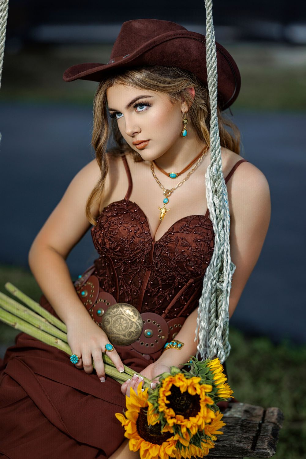 Young woman in brown corset dress and cowboy hat sits on rope swing holding sunflowers, wearing turquoise jewelry.