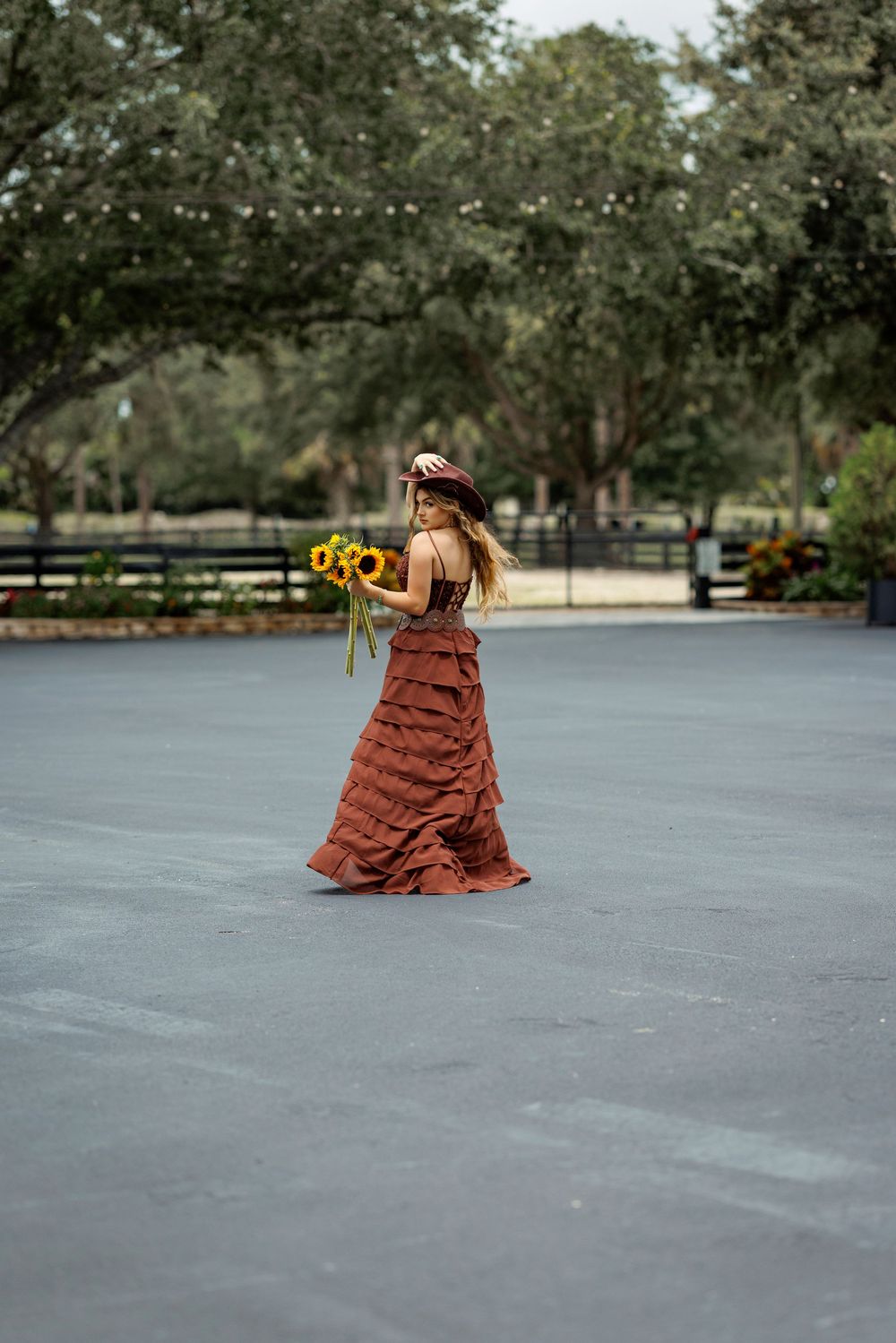 Woman in rust ruffled gown holding sunflowers walks across paved area with lush green trees backdrop.