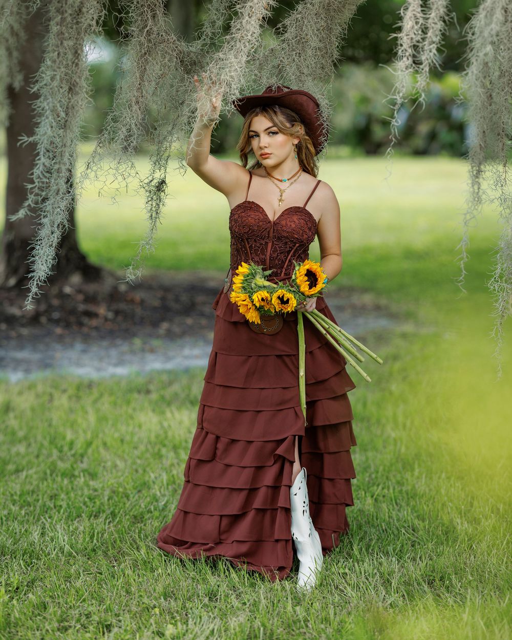Woman in brown tiered gown and hat holds sunflowers under Spanish moss-draped trees in a lush green setting.