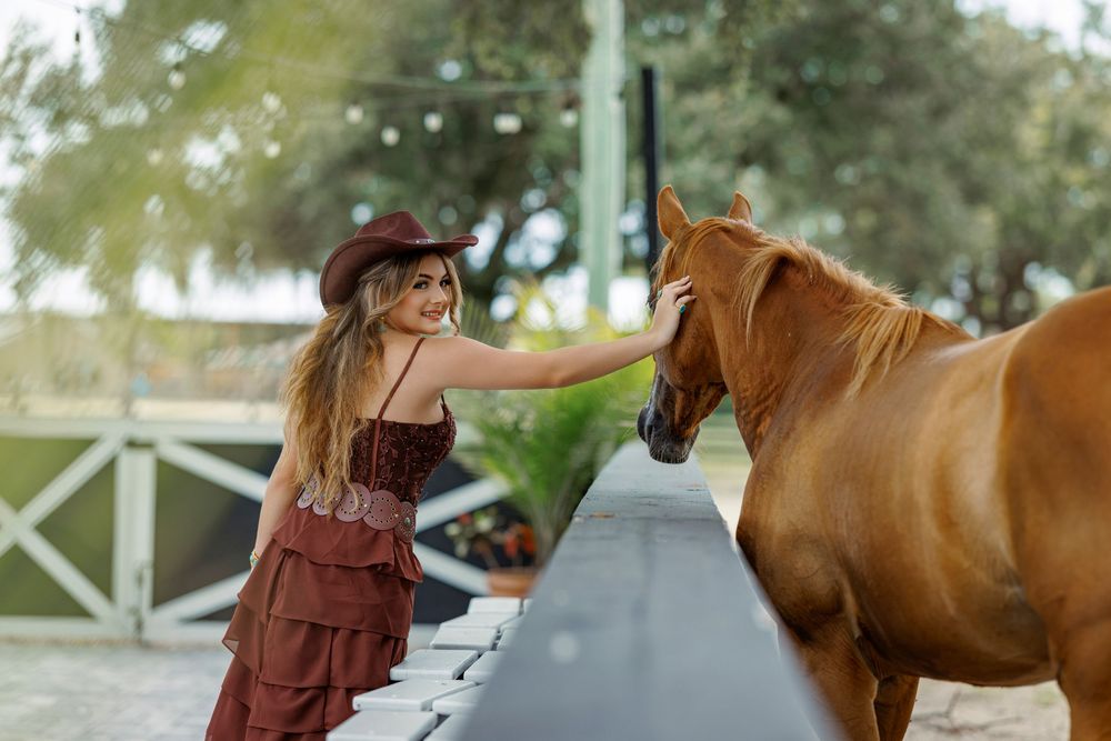 A smiling woman in a burgundy dress and cowboy hat gently pets a chestnut horse outdoors.