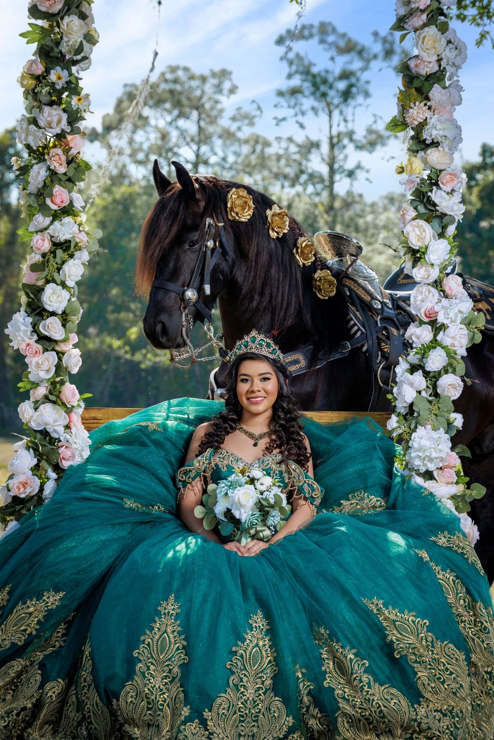 Smiling quinceañera in teal gold ballgown sits on floral swing beside decorated black horse.