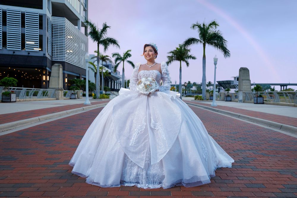 Bride in white ballgown poses on brick plaza with palm trees and rainbow at dusk.
