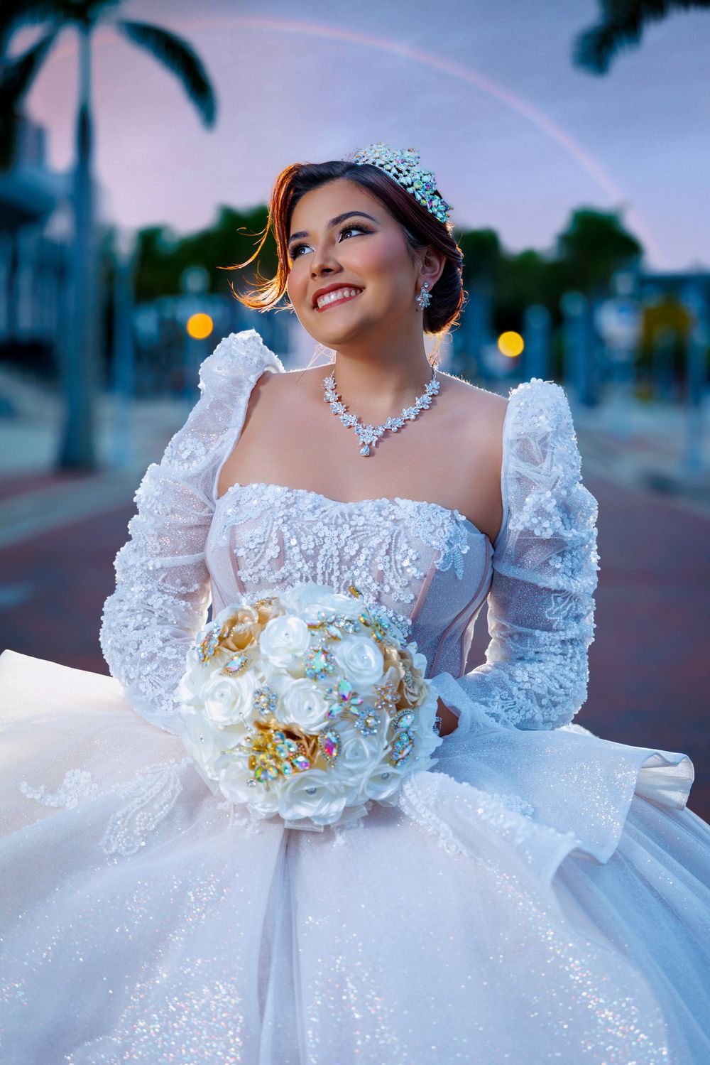 Smiling bride in white lace ballgown holds floral brooch bouquet with rainbow backdrop at dusk.