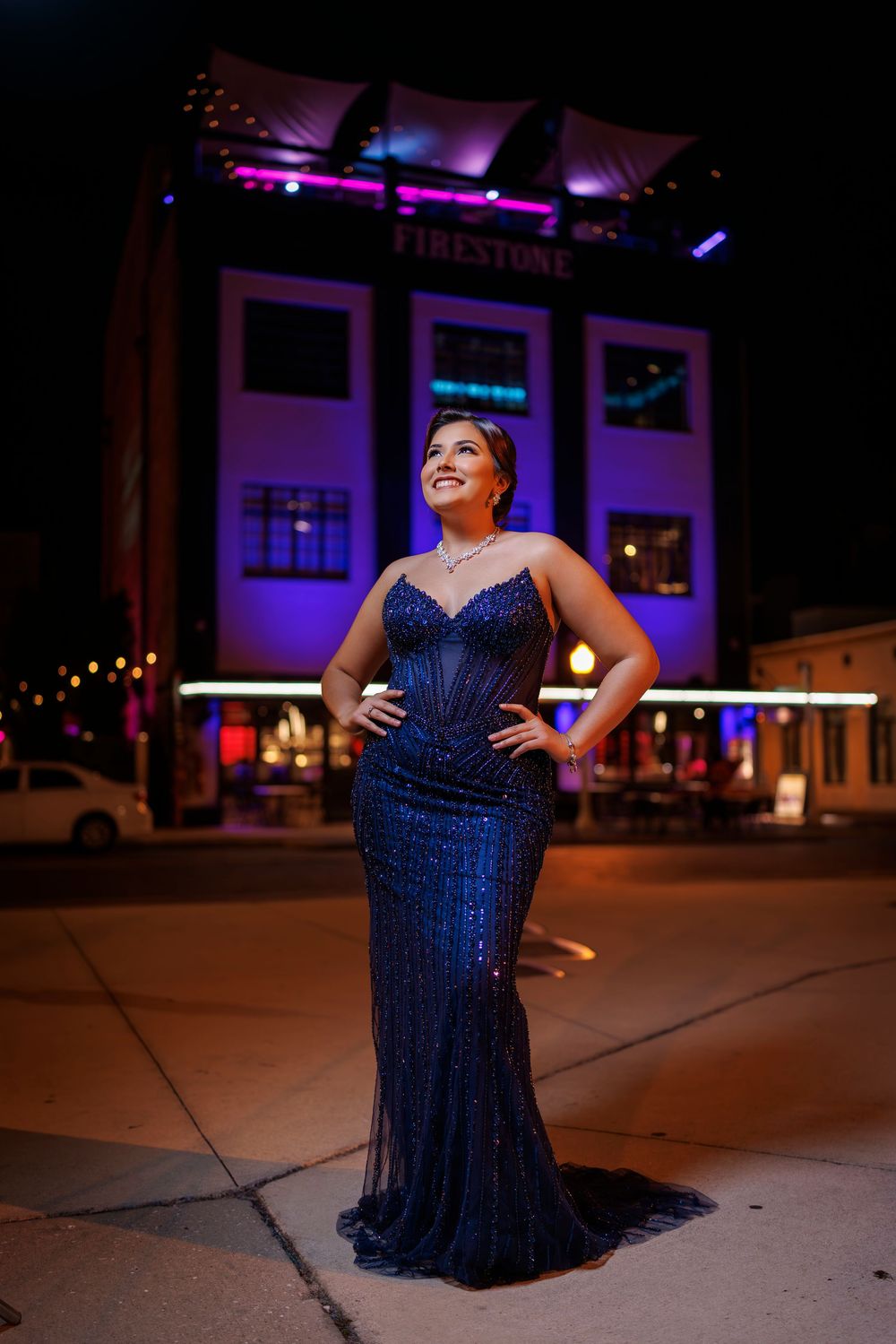 Woman in navy blue sequin mermaid gown smiling outside Firestone building at night.