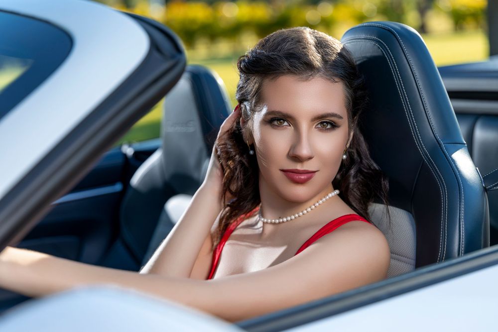 Young woman with pearl necklace in red dress sits in a white Porsche convertible smiling at camera.