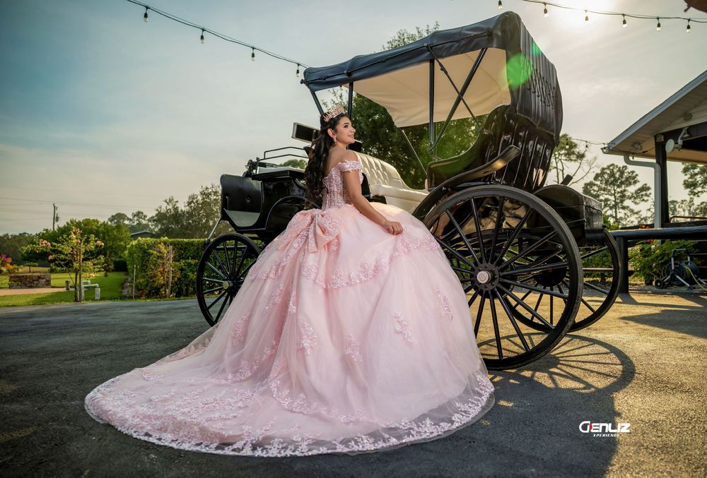 Quinceañera girl in pink ball gown sits beside elegant black horse-drawn carriage outdoors.