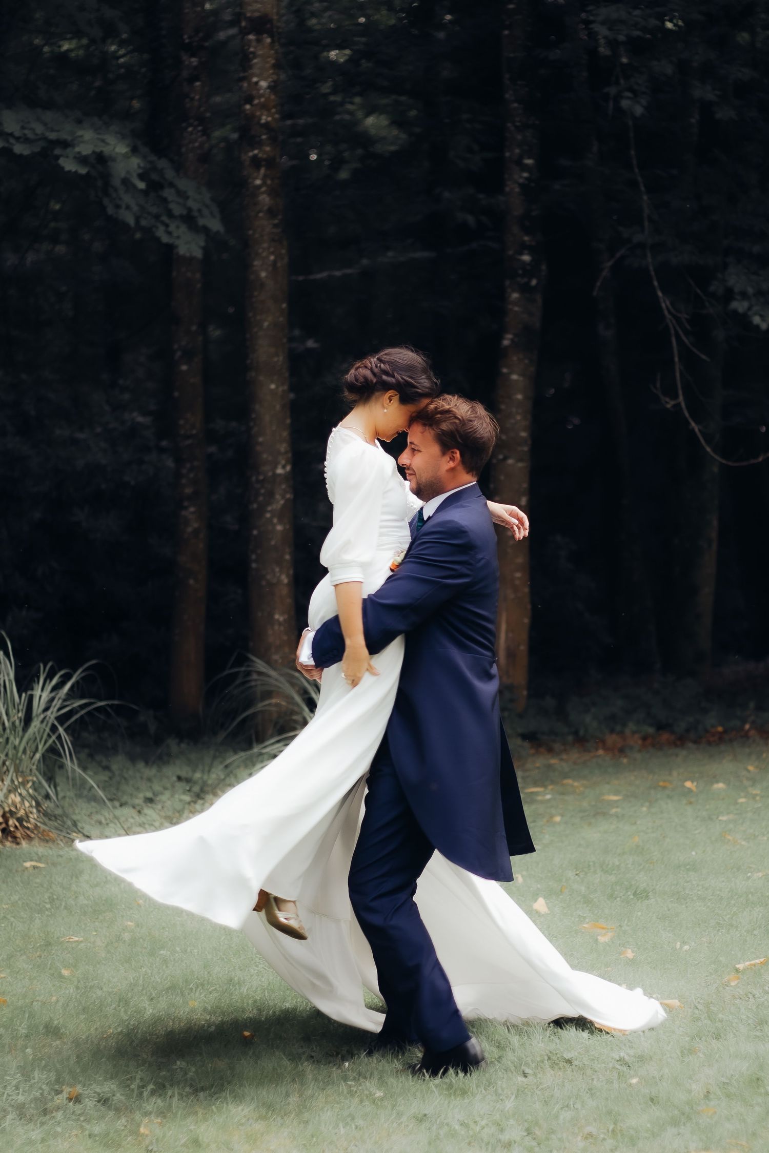 Groom in navy suit lifting and spinning bride in white dress in a lush forest setting.