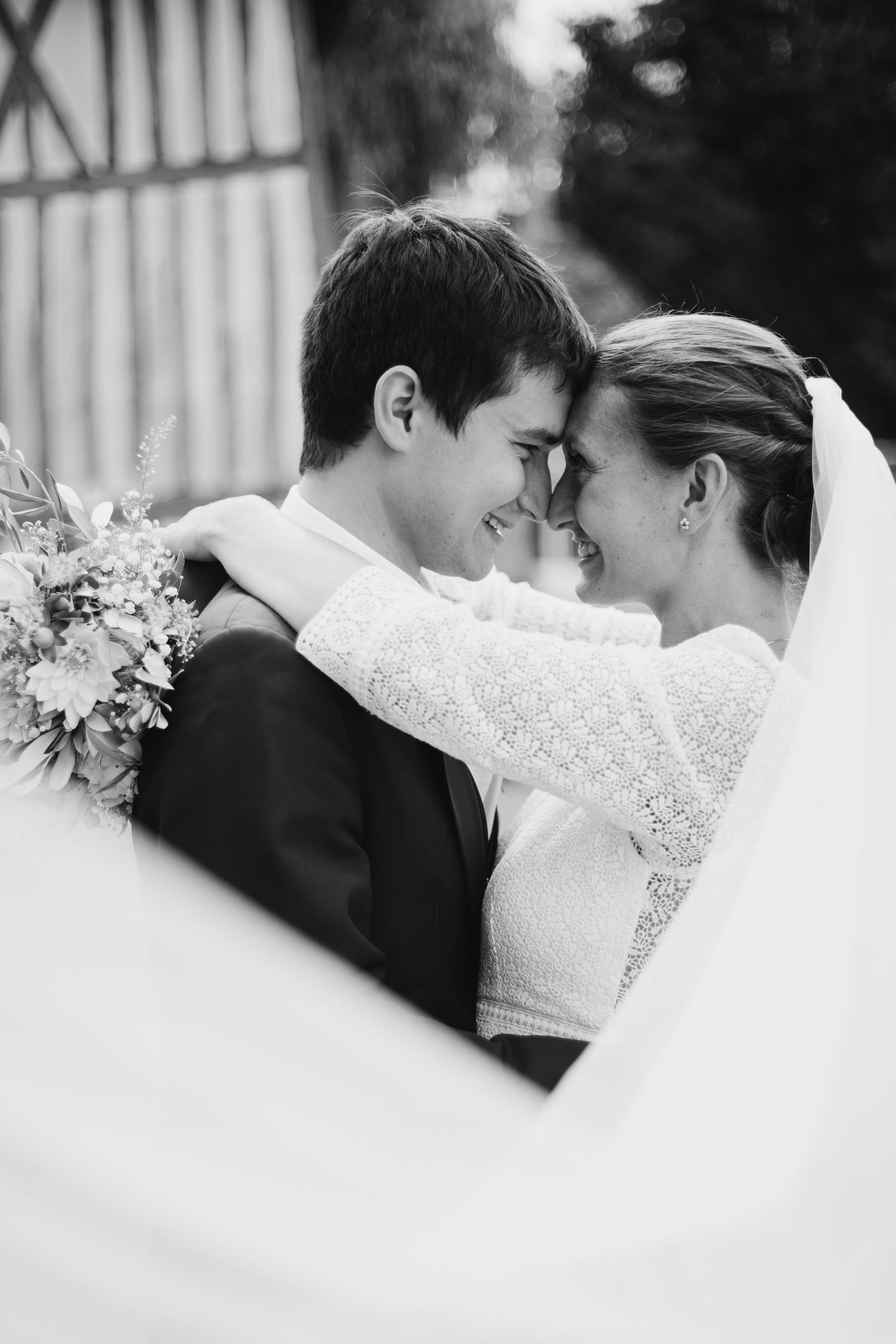 Black and white photo of smiling bride and groom touching foreheads, bride holding bouquet with veil flowing.