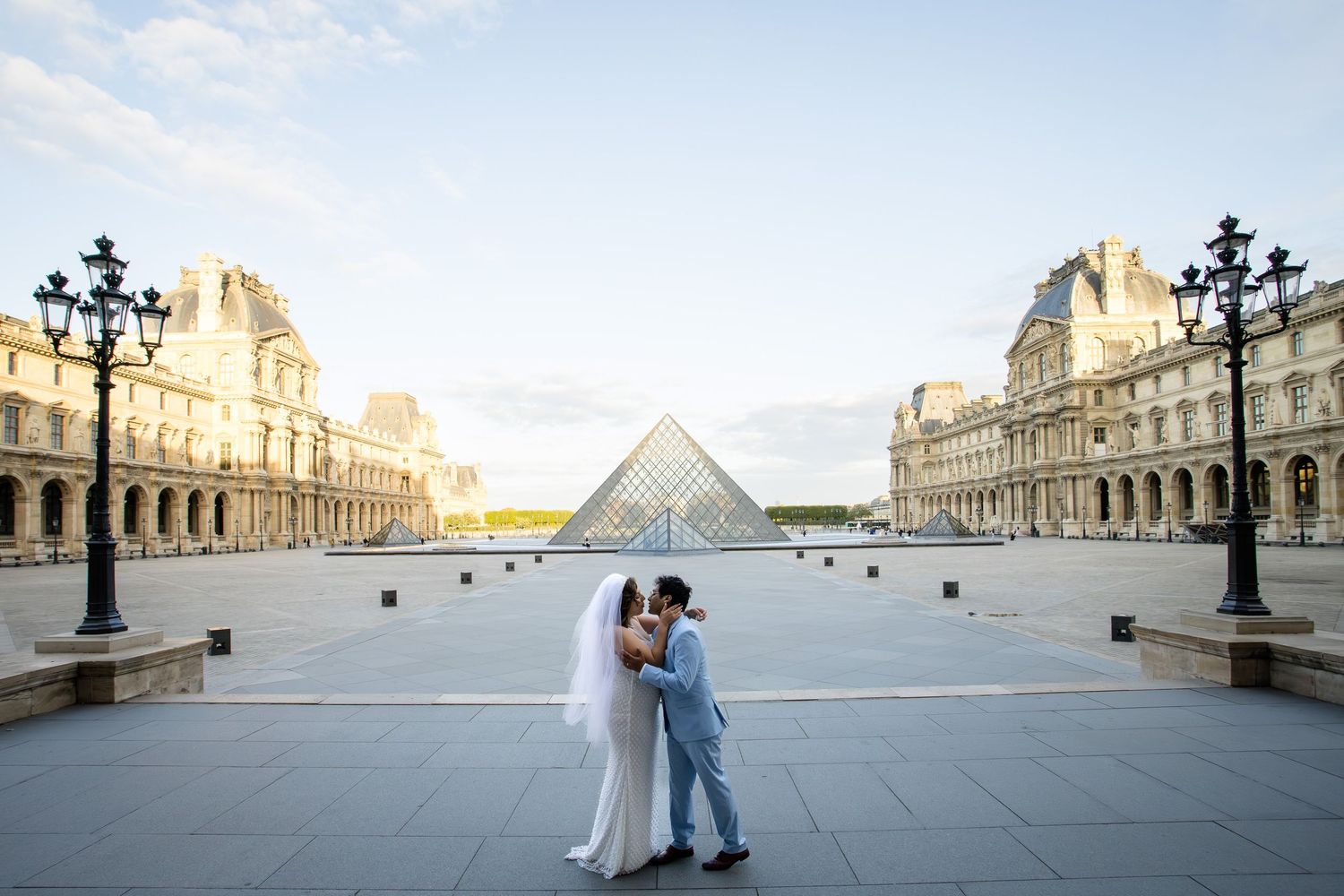 A romantic couple in blue and white outfits embrace and dance in front of the iconic Louvre Museum in Paris.