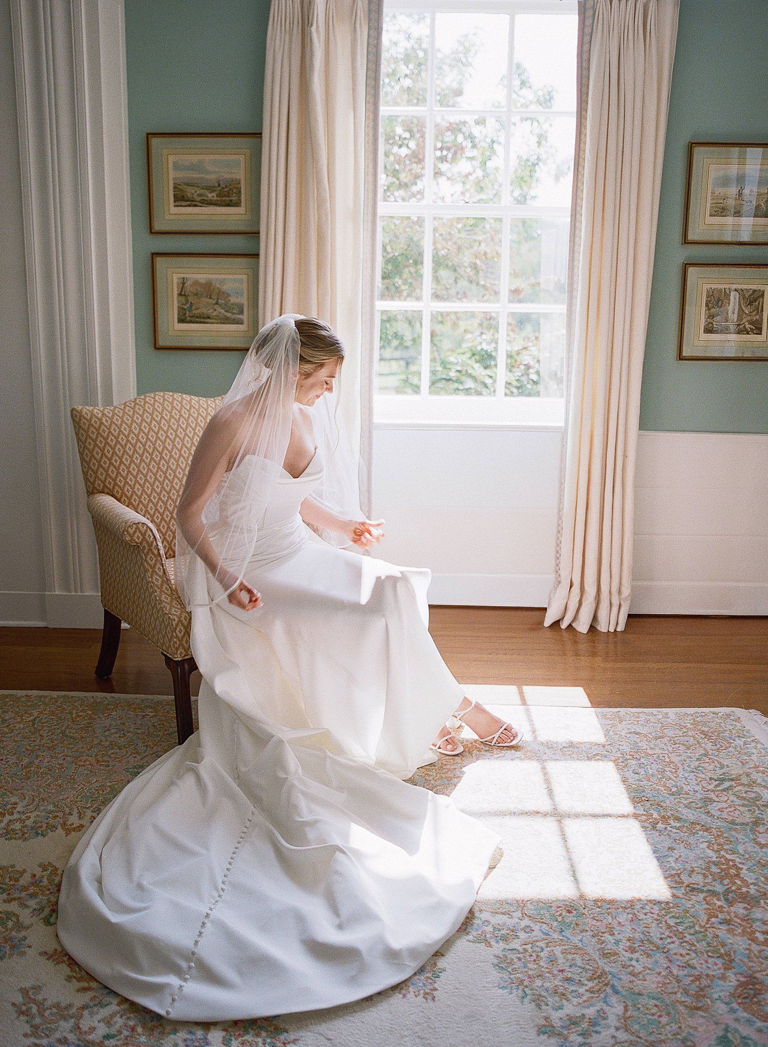 A flowing white wedding dress swirls gracefully across a sunlit room with floral carpeting and sage green walls.
