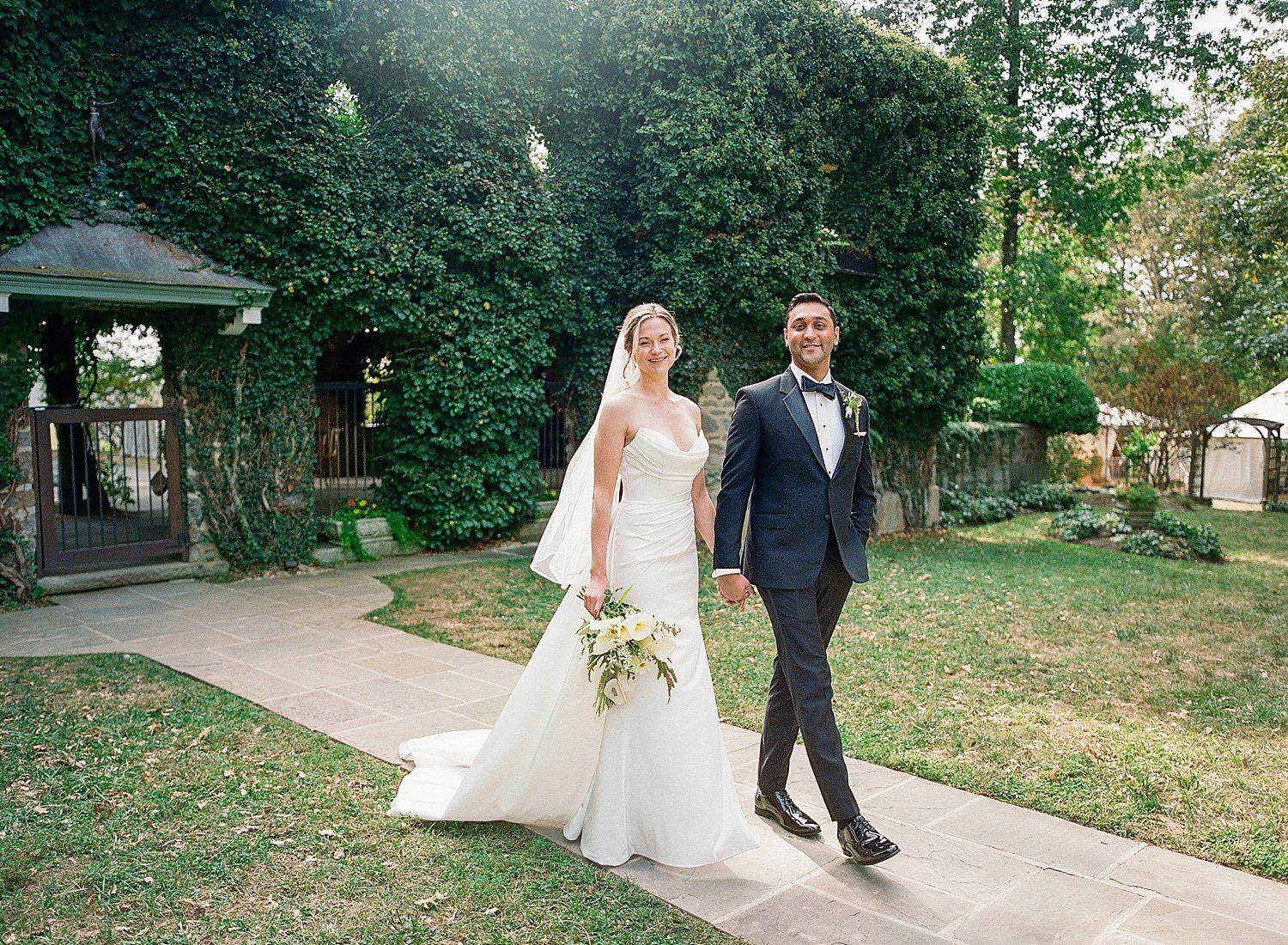 A couple walks along a garden path surrounded by lush greenery on their wedding day.