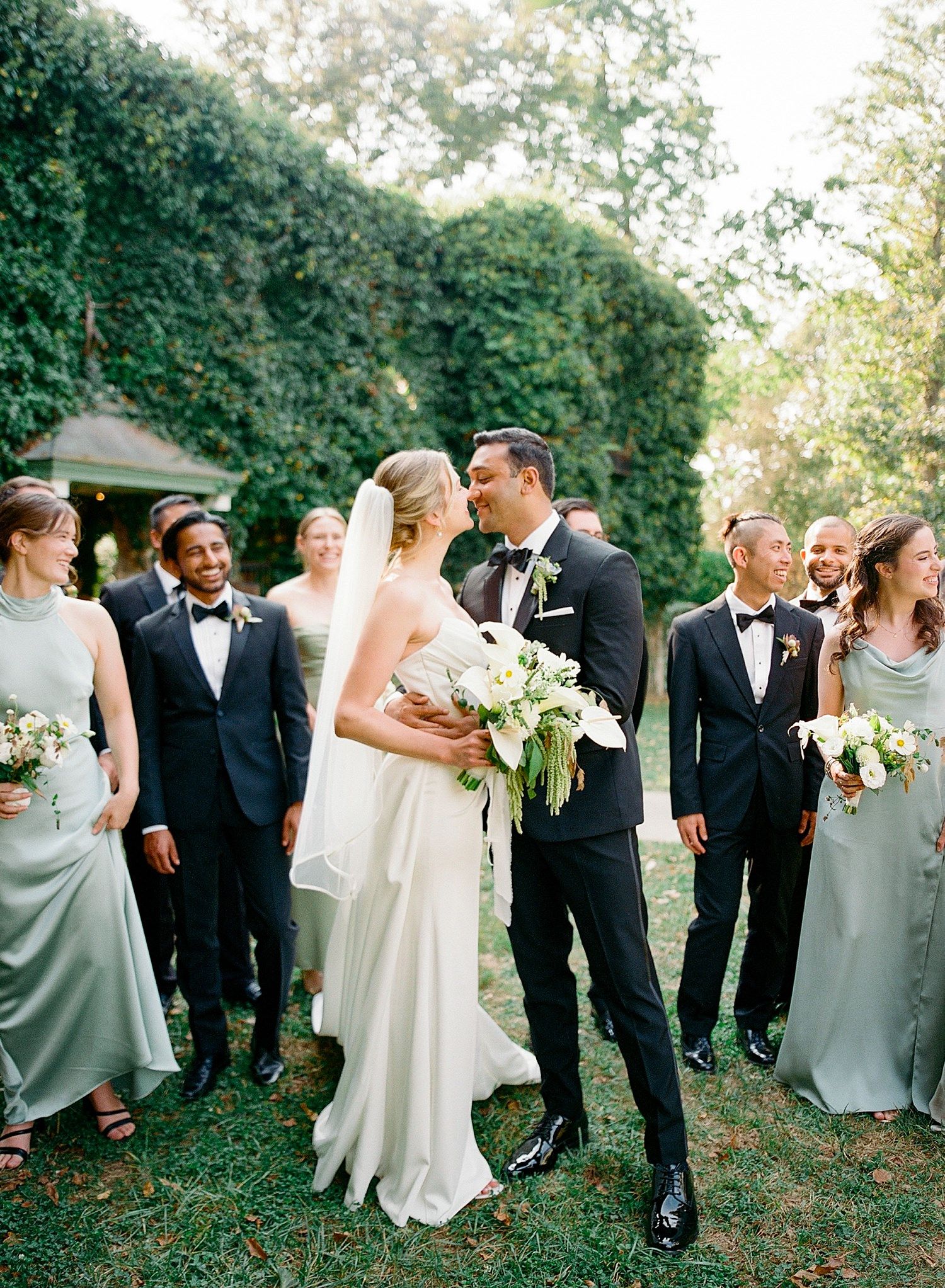 Wedding party celebrates outdoors with bridesmaids in sage green dresses and groomsmen in black tuxedos during garden ceremony.