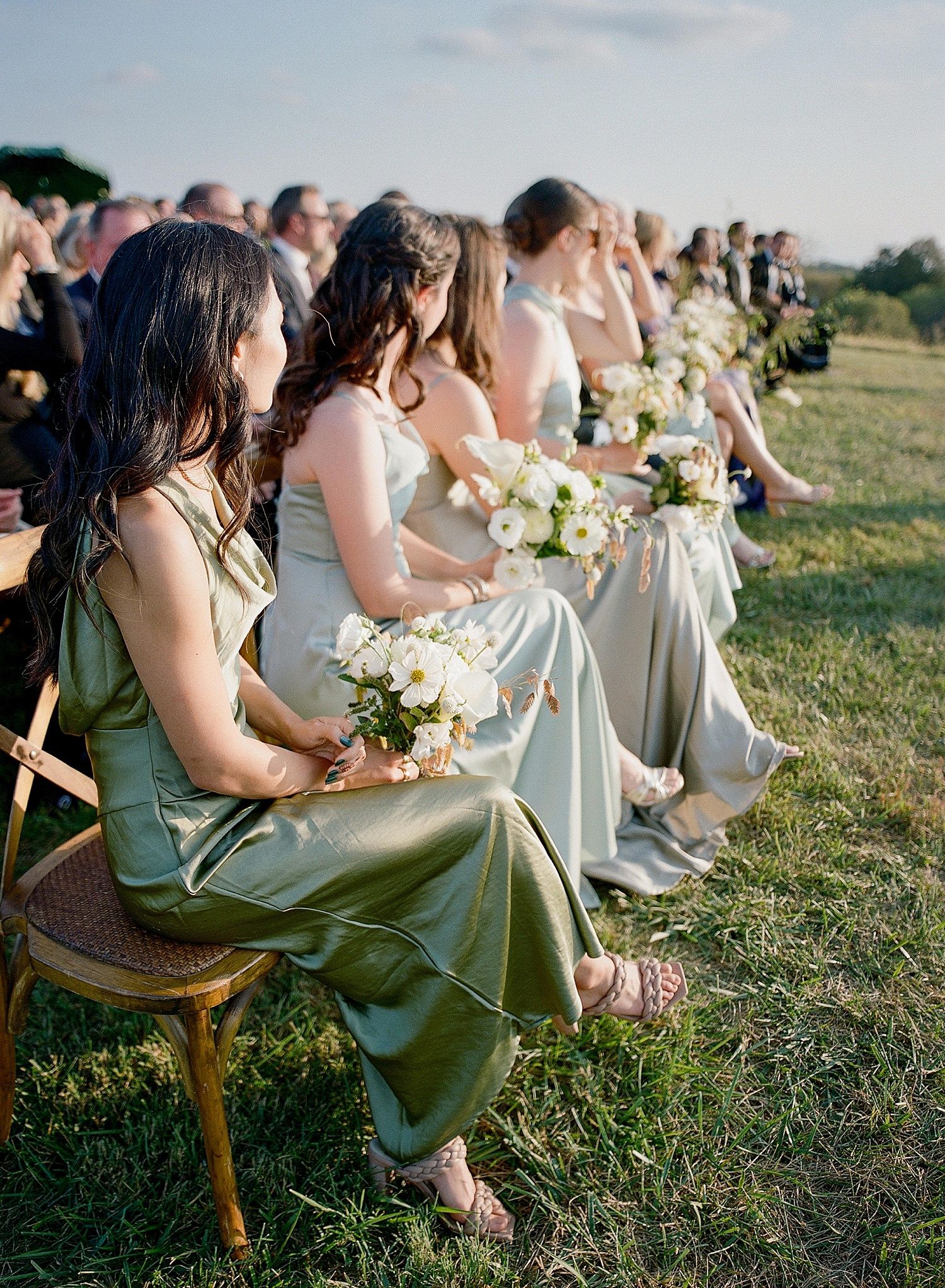 Wedding guests in sage green dresses sit on wooden chairs outdoors holding floral bouquets during ceremony.
