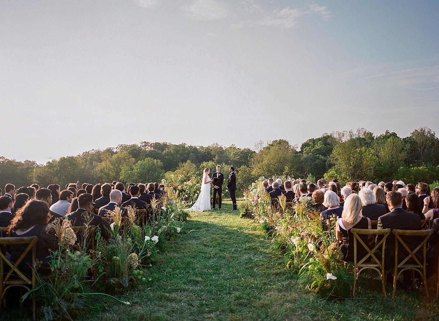 Panoramic view of an outdoor wedding ceremony set in a lush green hillside meadow with wooden cross-back chairs.