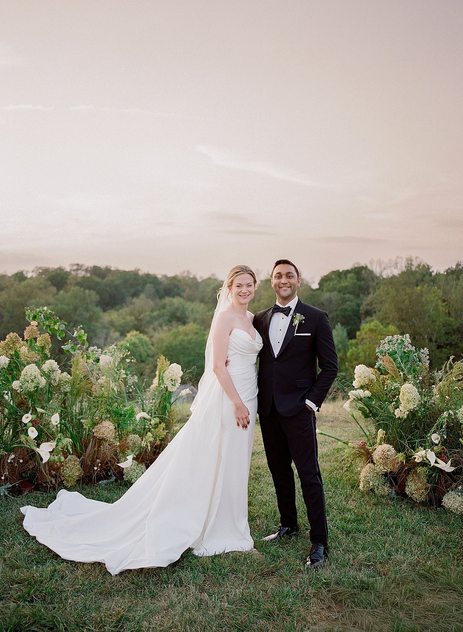 A couple stands together in a romantic outdoor garden setting with white hydrangeas at sunset.